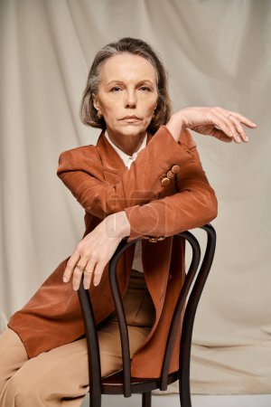 Foto de Una mujer madura y atractiva con una chaqueta marrón se sienta graciosamente en una silla, exudando una sensación de paz y elegancia. - Imagen libre de derechos