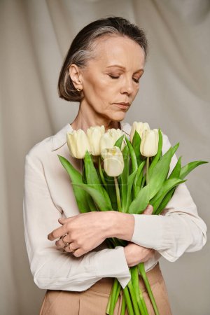 A mature woman in comfy attire gracefully holds a bouquet of white tulips.