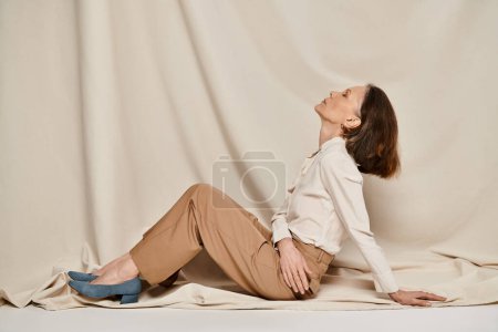 Eine Frau in weißem Hemd und brauner Hose sitzt anmutig auf einem Bett.