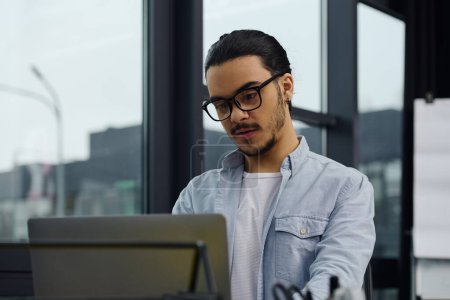 Photo for A man in glasses focusing on work on a laptop in an office setting. - Royalty Free Image