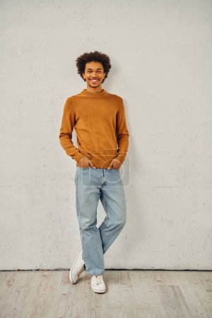 Foto de Hombre joven en jersey bronceado y jeans de pie con una expresión reflexiva contra una pared. - Imagen libre de derechos