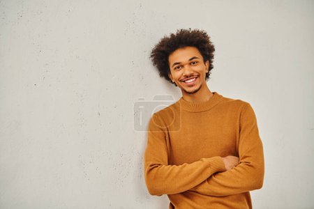 Foto de Un joven con estilo en un suéter bronceado apoyado casualmente contra una pared. - Imagen libre de derechos