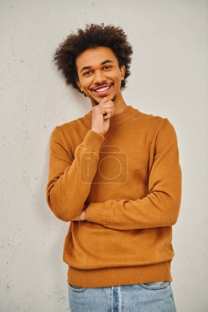 Un homme en pull marron appuyé contre un mur.