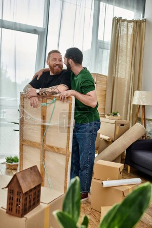 Deux hommes se tiennent ensemble dans un salon rempli de boîtes mobiles, commençant leur vie ensemble dans une nouvelle maison.
