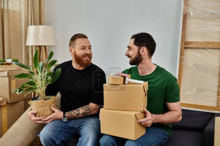 A gay couple in love, holding boxes and a plant, prepares for a new life in their new home filled with moving boxes.