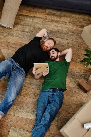 Foto de A loving gay couple laying tenderly on a wooden floor amid moving boxes in their new home. - Imagen libre de derechos