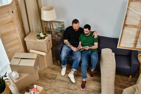 A gay couple sit atop a couch, embracing in their new home among boxes, symbolizing love and new beginnings.