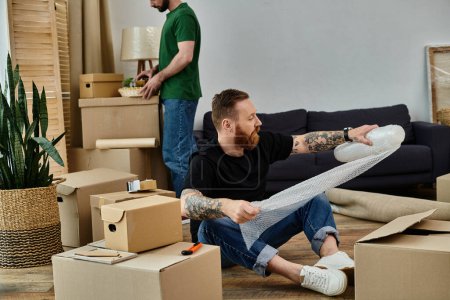 Téléchargez les photos : A man sits surrounded by moving boxes, embracing change as a gay couple starts a new life in a new home. - en image libre de droit