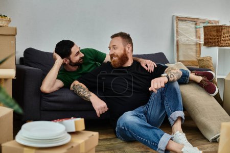A gay couple relaxes together atop a couch in their new home, surrounded by unpacked boxes, marking the start of a new chapter.