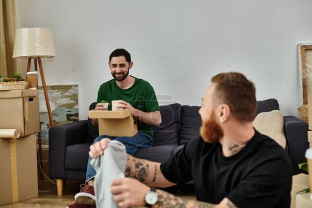 A man, part of a gay couple in love, sits on a couch holding a box as they move into their new home filled with boxes.