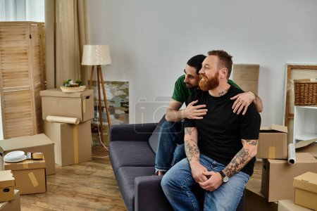 Foto de A gay couple in love sit on top of a couch in their new home, surrounded by moving boxes, embracing their new life. - Imagen libre de derechos