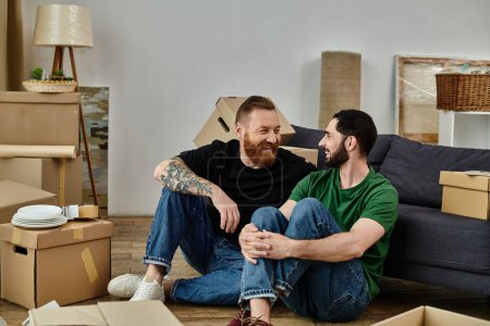 Foto de Gay couple sitting on wooden floor, surrounded by moving boxes, starting new life together in cozy home. - Imagen libre de derechos