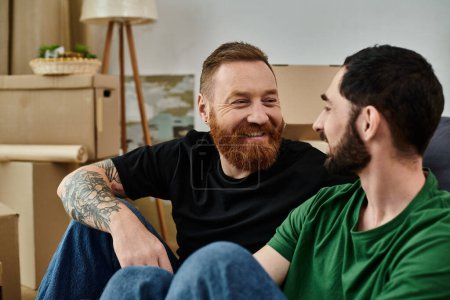 Photo for Two men, part of a gay couple in love, relax on a couch in their new home surrounded by moving boxes. - Royalty Free Image