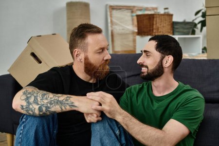 Two men, part of a gay couple, sit happily atop a couch in their new home, amidst moving boxes and the promise of a fresh start.