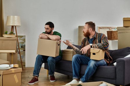 Photo for Gay couple sitting on sofa surrounded by moving boxes in their new home, having disagreement and misunderstanding - Royalty Free Image