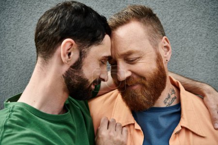Foto de Una pareja gay cariñosa con atuendo casual se paran lado a lado contra una pared gris, exudando unidad y afecto. - Imagen libre de derechos