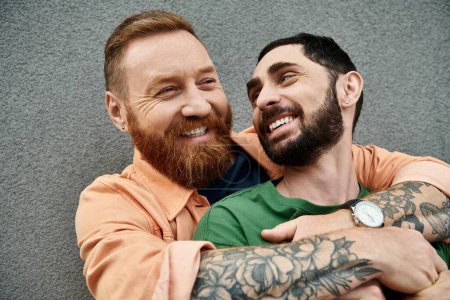 Foto de Two men with tattoos on arms, in casual attire, embrace in love, standing against grey wall. - Imagen libre de derechos