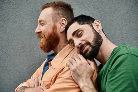 Schwules Paar in lässiger Kleidung, zwei Männer mit Bärten, umarmen einander vor einer grauen Wand in einer Zurschaustellung von Liebe und Einheit.