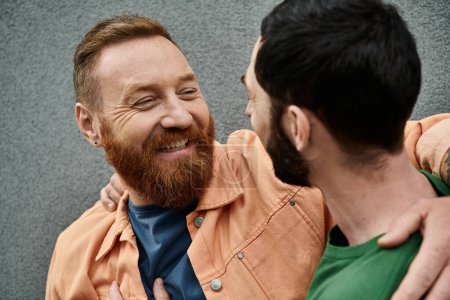 A bearded man embraces another man lovingly against a grey wall, both in casual attire.