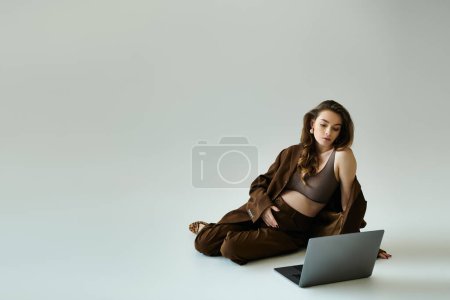Eine junge schwangere Frau im braunen Anzug mit Blazer sitzt auf dem Boden, während sie an einem Laptop arbeitet.