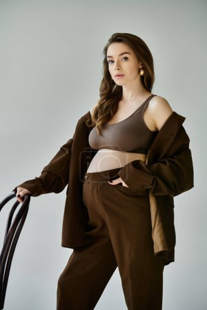 A young pregnant woman elegantly poses in a stylish brown suit and blazer against a soft grey backdrop.