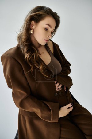 Young pregnant woman in a stylish brown coat poses gracefully for a portrait on a grey background.