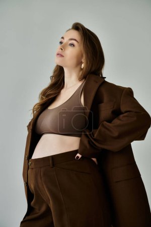 Foto de Una joven embarazada con estilo en una chaqueta marrón y pantalones se levanta con gracia sobre un fondo gris. - Imagen libre de derechos