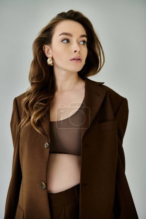 A young pregnant woman exudes elegance in a brown jacket on a grey background.