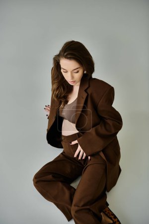 Photo for Pregnant woman in brown suit with blazer sits on ground, deep in thought, against grey backdrop. - Royalty Free Image