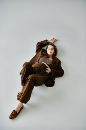 A serene young pregnant woman in a sophisticated brown outfit lounging gracefully on the ground.