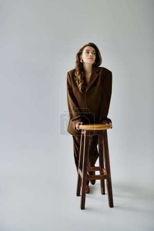 A young pregnant woman in a brown suit with a blazer leans gracefully on top of a wooden stool against a grey background.