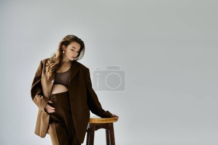 Eine junge schwangere Frau im braunen Anzug steht anmutig auf einem Holzhocker vor grauem Hintergrund..