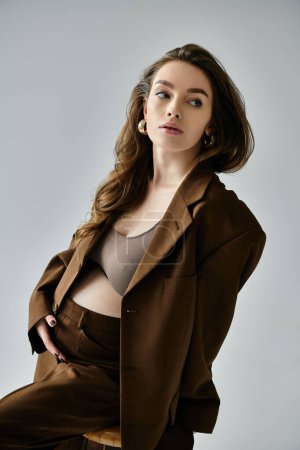 A young pregnant woman confidently poses in a stylish brown suit with a blazer against a neutral grey backdrop.