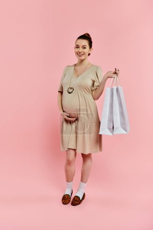 Eine junge schwangere Frau in einem Kleid hält freudig eine Einkaufstasche in der Hand, während sie auf rosa Hintergrund lächelt.