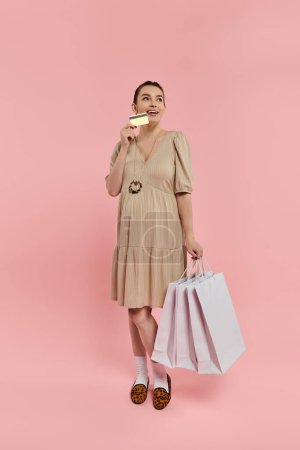 A stylish young pregnant woman in a dress balancing two shopping bags and a credit card on a pink background.