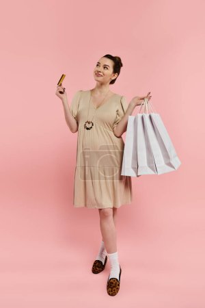 Eine schwangere Frau in einem Kleid hält zwei Taschen und eine Kreditkarte vor einem leuchtend rosa Hintergrund.