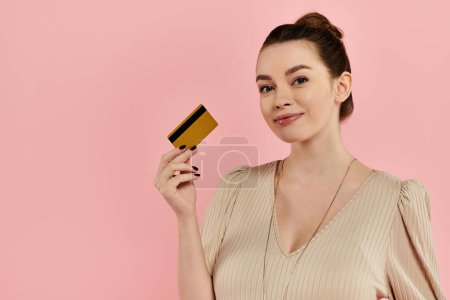 Une femme enceinte tient élégamment une carte de crédit dans sa main sur un fond rose.