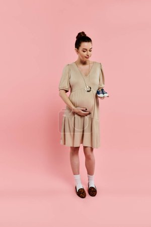 A pregnant woman in a dress lovingly holds a small baby shoe against a soft pink background.