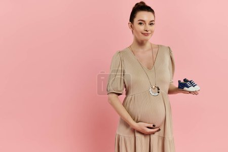 A young pregnant woman in a dress gracefully holds shoes against a soft pink backdrop.