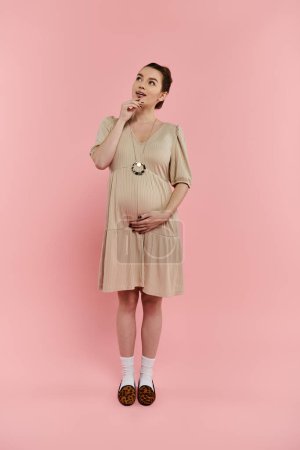 Junge schwangere Frau steht selbstbewusst in einem fließenden Kleid vor einem leuchtend rosa Hintergrund.