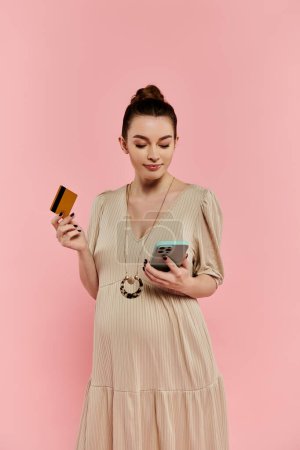 Una joven embarazada en un vestido con un teléfono celular y una tarjeta de crédito sobre un fondo rosa.