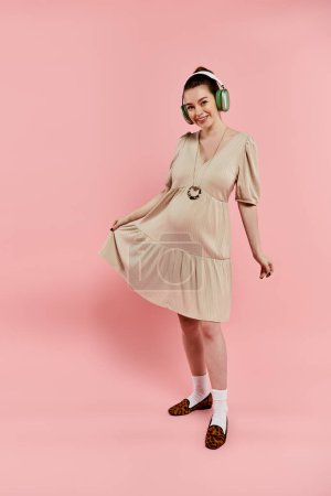 Young pregnant woman in a flowing dress, wearing headphones, striking a pose for a portrait on a pink background.