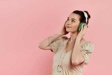 Young pregnant woman in a dress joyfully listening to headphones on a pink background.
