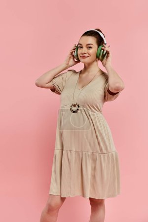A stylish pregnant woman in a flowing dress listens to music through headphones against a vibrant pink backdrop.