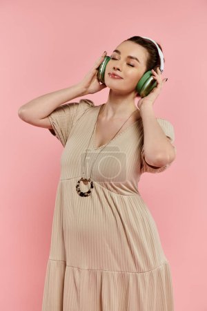 A young pregnant woman in a dress listening to headphones on a pink background.