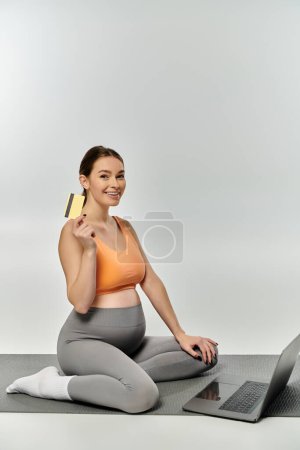 A pregnant woman in active wear holds a credit card on a yoga mat, embodying financial wellness and balance.
