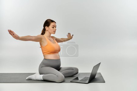 Eine junge schwangere Frau sitzt auf einer Yogamatte und benutzt einen Laptop zum Arbeiten und Entspannen.