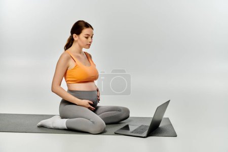 Foto de Una mujer embarazada deportiva se sienta en una esterilla de yoga, multitarea con un ordenador portátil. - Imagen libre de derechos