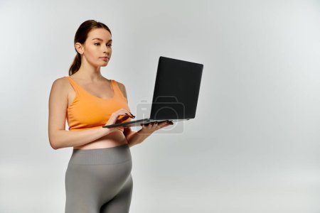 Eine junge, sportliche Schwangere in aktiver Kleidung hält einen Laptop in der Hand.