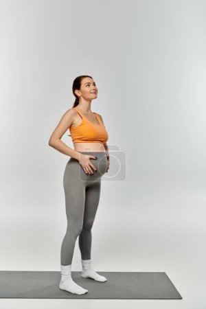 Mujer embarazada joven en desgaste activo de pie sobre estera de yoga sobre fondo gris.